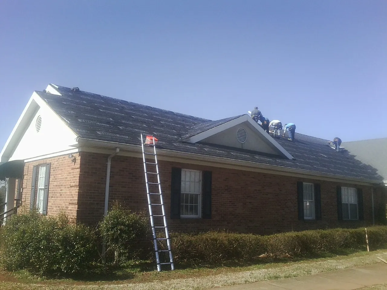 Crew tearing shingles off a brick ranch roof with a ladder set — Emergency Roof Repair work by our crew in Tar Heel
