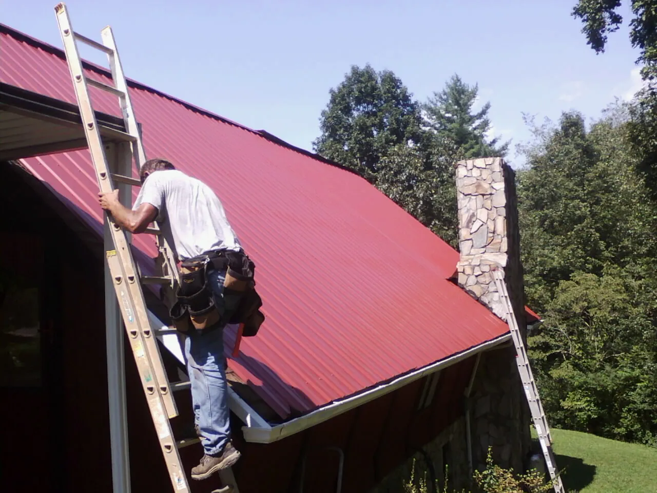 Roofer on a ladder beside a red metal roof and stone chimney — Soffit & Fascia Repair by Roofing Brothers in Tar Heel